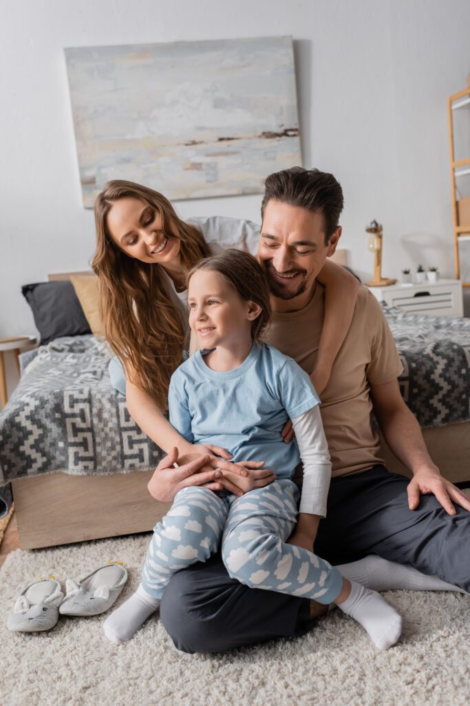 cheerful kid sitting with positive father on carpet near happy mother on bed.
