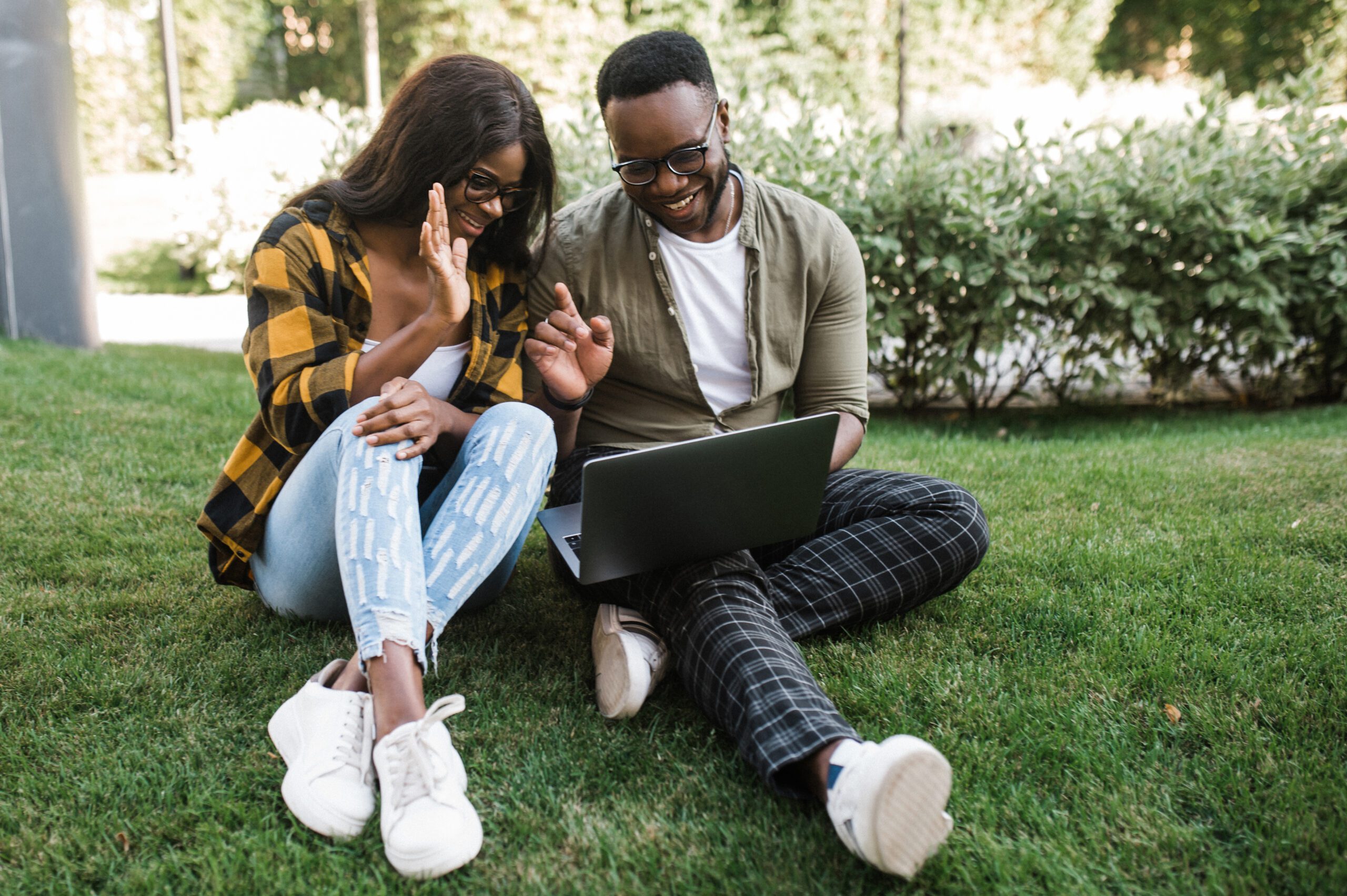 Young African American guy and girl dressed in stylish casual clothes are sitting on the grass with a laptop and chatting with friends or colleagues via video conference