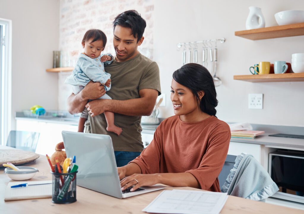 A woman works on a laptop at a kitchen table, smiling. A man stands beside her, holding a baby. The scene conveys a warm, family-oriented atmosphere.
