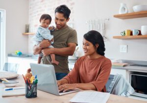 A woman works on a laptop at a kitchen table, smiling. A man stands beside her, holding a baby. The scene conveys a warm, family-oriented atmosphere.