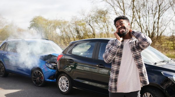A man stands beside two crashed cars on a rural road, holding his neck and phone, looking concerned. The front car is black, and the rear blue car is smoking.